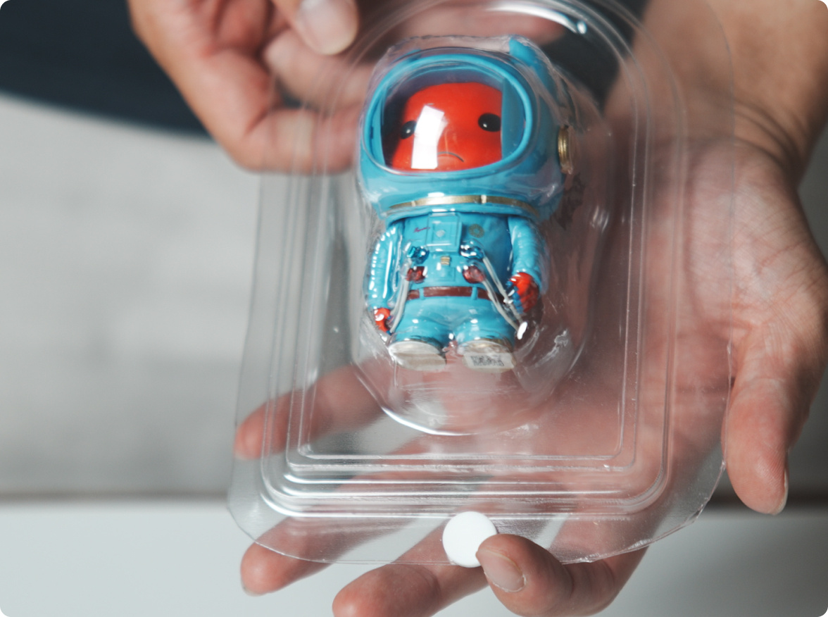 Two hands holding a clamshell blister containing a figurine of a cute astronaut with red skin wearing a teal suit