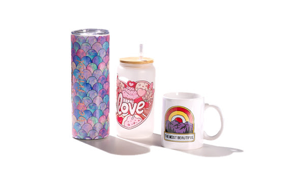 Stainless steel tumbler with sublimated colorful shell motif, glass mug with lid and straw and sublimated heart shape and love text, sublimated ceramic mug with red and yellow rainbow, mountain range and 'the most beautiful' in text.