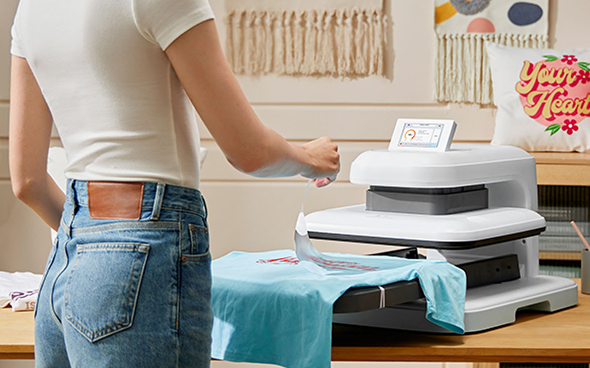 LOKLiK ImPress™ Auto 2-Smart in Daisy White on a wooden table. A woman is peeling of a transfer sheet from a blue shirt that is lying on the ironing board.