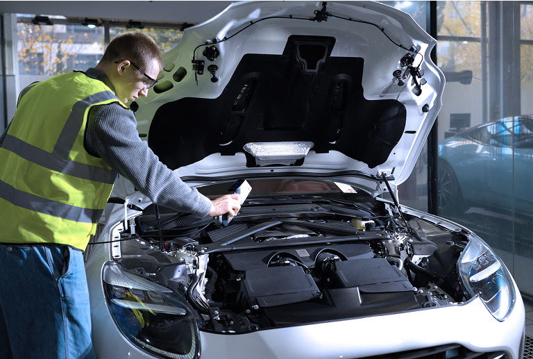 Man in a safety vest bending over a car with the hood open. He is scanning the enginge part of the car. 