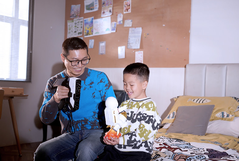 A man and a young boy sitting on a bed. The man is holding the 3DeVOK MQ with the wireless handle and he is 3D scanning an astronaut figurine the boy is holding.