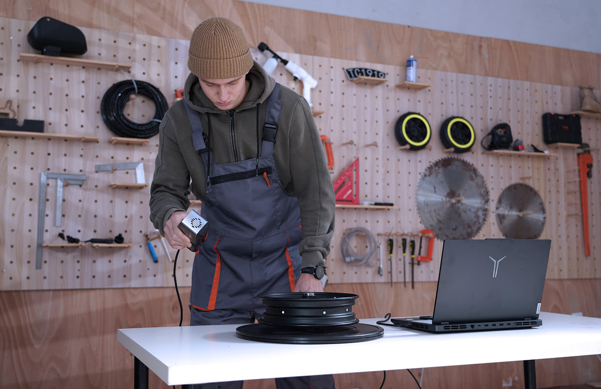 Man in a car workshop 3D scanning a car rim  on a table. Next to it on the table is an open laptop.
