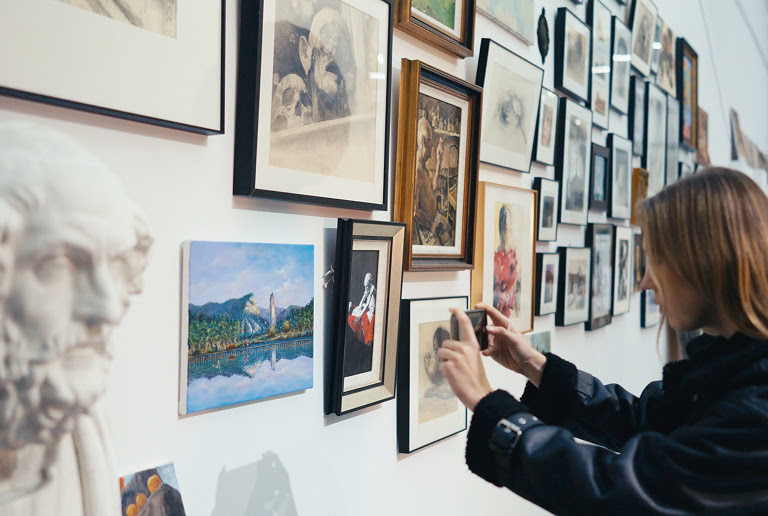 Woman taking a photo of a picture on a wall with several pictureframes.