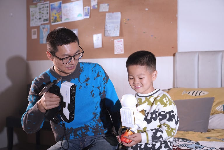 A man and a young boy sitting on a bed. The man is holding the 3DeVOK MQ with the wireless handle and he is 3D scanning an astronaut figurine the boy is holding.