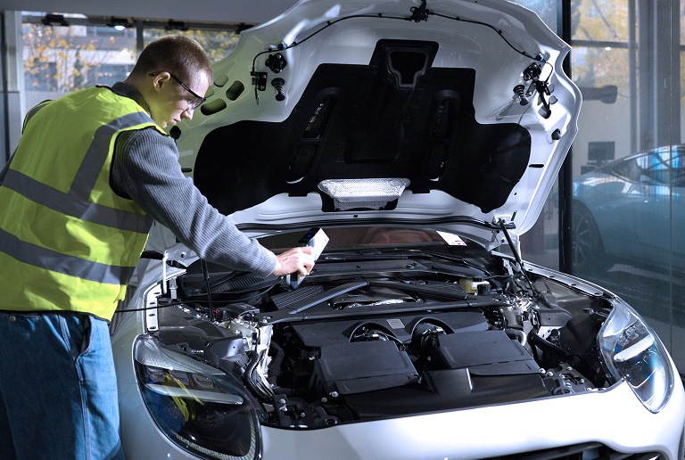 Man in a safety vest bending over a car with the hood open. He is scanning the enginge part of the car. 