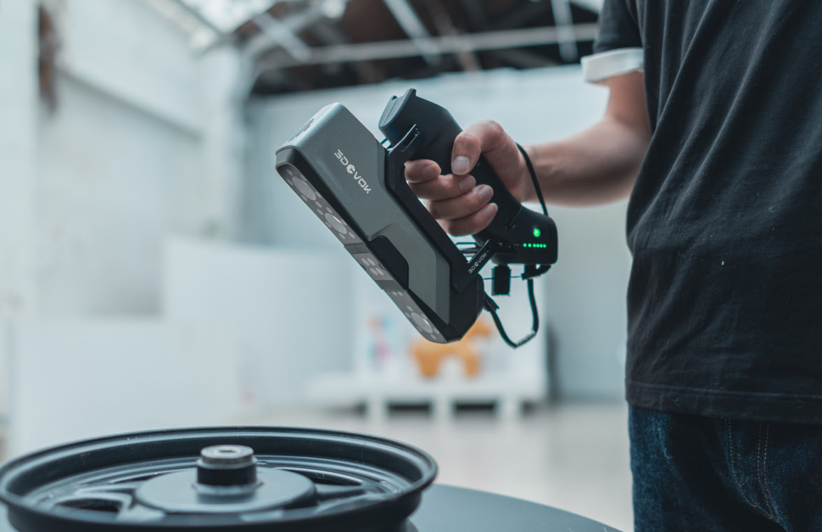 Hand holding a 3DeVOK MT with a wireless handle attached to it. The person is scanning a black car wheel.