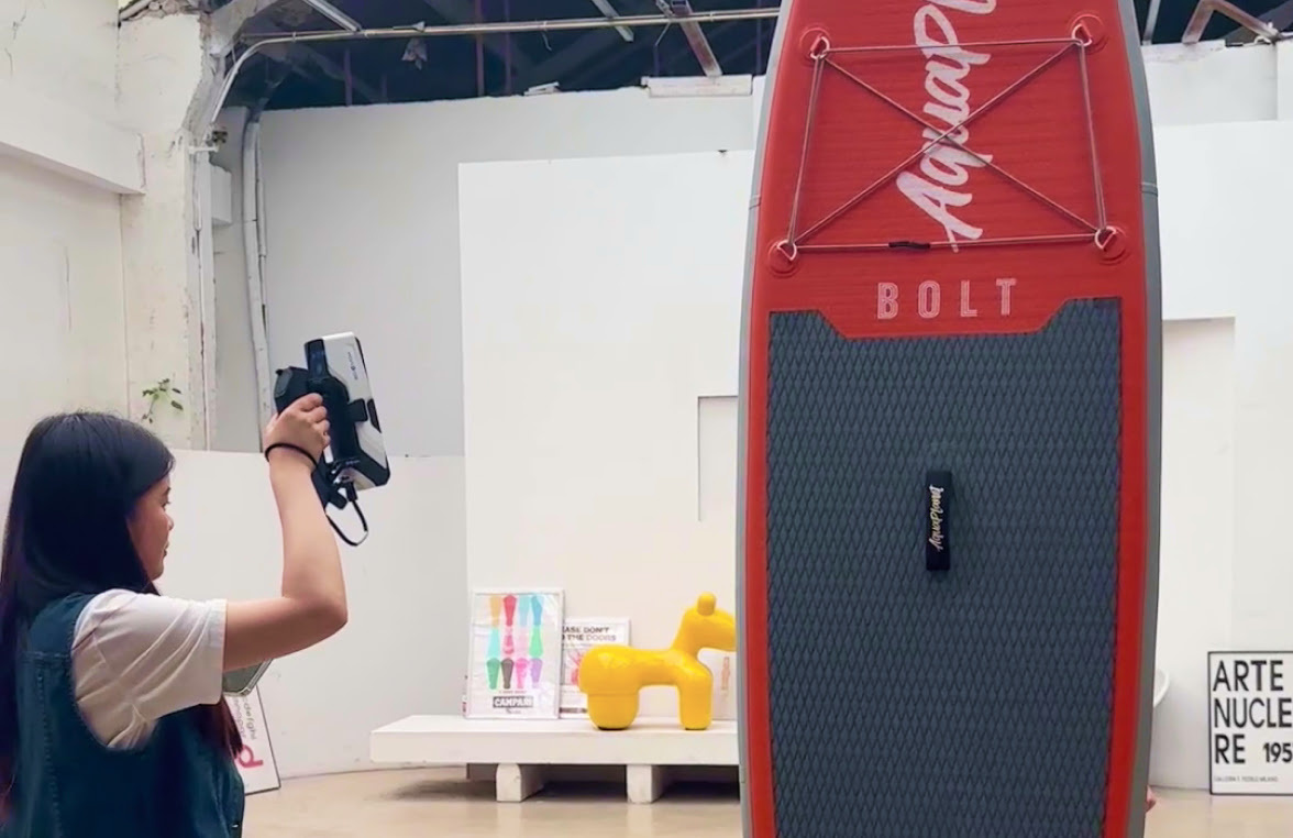 Woman holding a 3DeVOK MQ with a wirelesshandle. She as raised the 3D scanner above her head and is 3D scanning a red surfboard.
