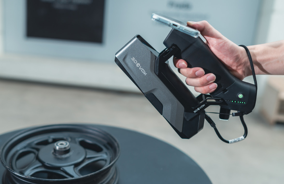 Hand holding a 3DeVOK MT with a wireless handle attached to it. The person is scanning a black car wheel.
