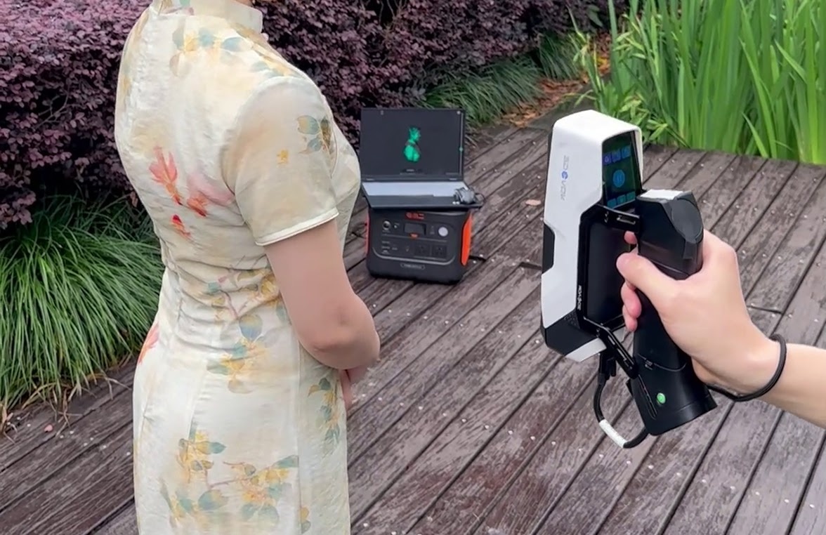 Hand holding a 3DeVOK MQ with a wireless handle. A person in a dress with a flower print is being scanned. They are outside.