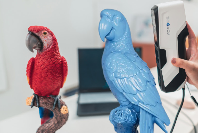 Hand holding a 3DeVOK MQ while scanning a colourful red parrot. There is a blue render of the same parrot imposed on the picture.