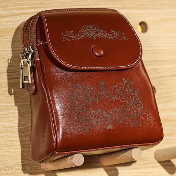 Leather satchel resting on two wooden pegs. It has an intricate design with swirls engraved on it.
