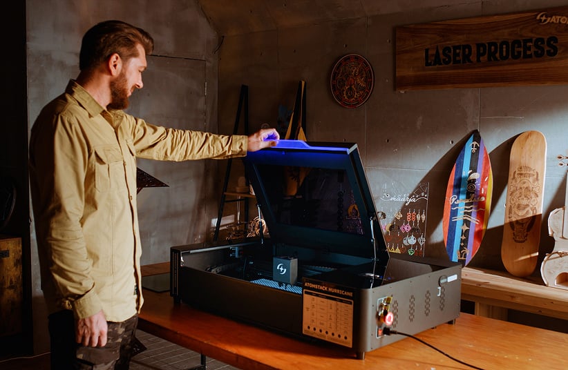 Man in a workshop opening an AtomStack Kraft on a wooden table. The blue led light of the Hurricane is on.