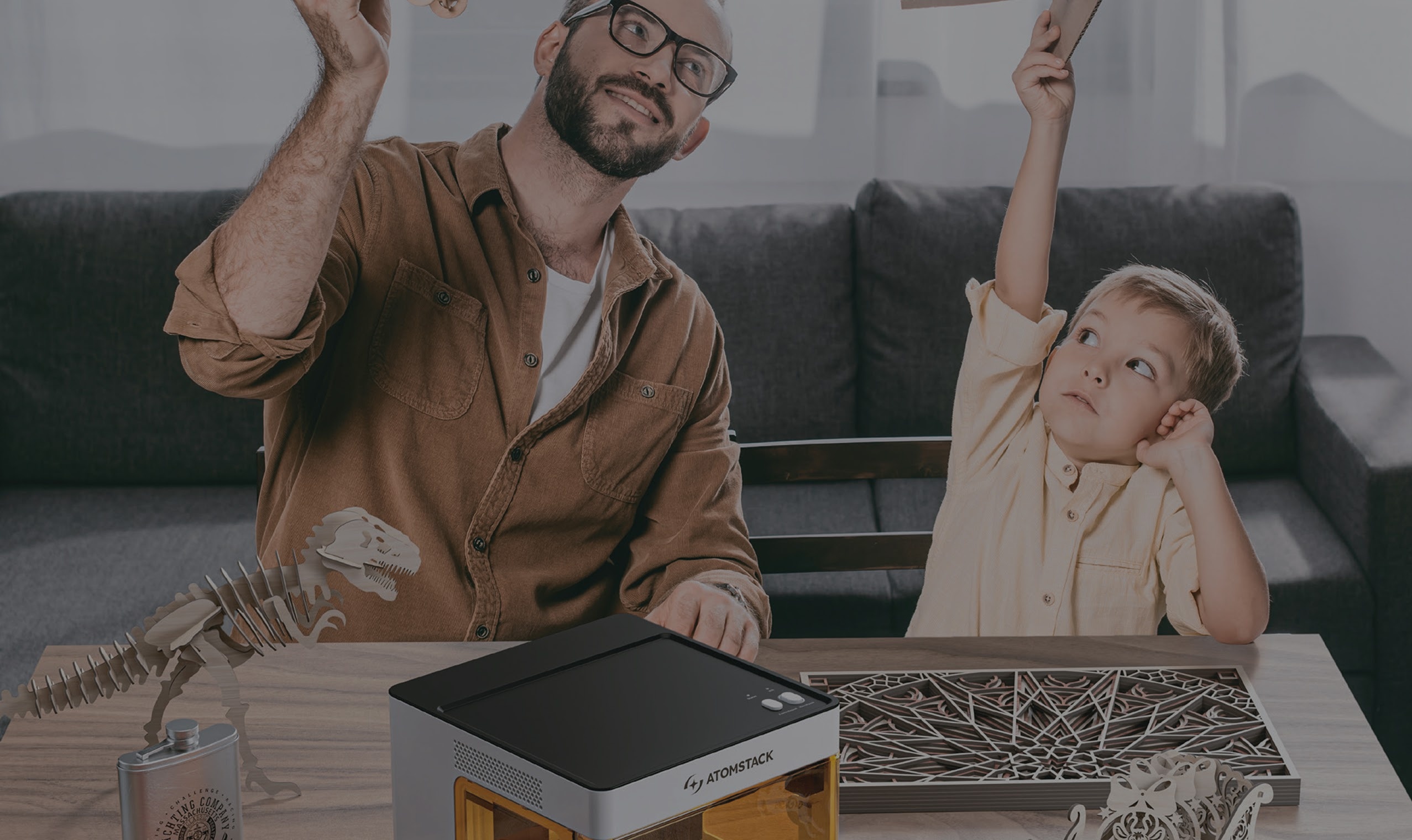 Man and younger child sitting at a low table raising their hand in the air. They are holding a laser cut airplane. On the table in front of them is the AtomStack P1 laser engraver.
