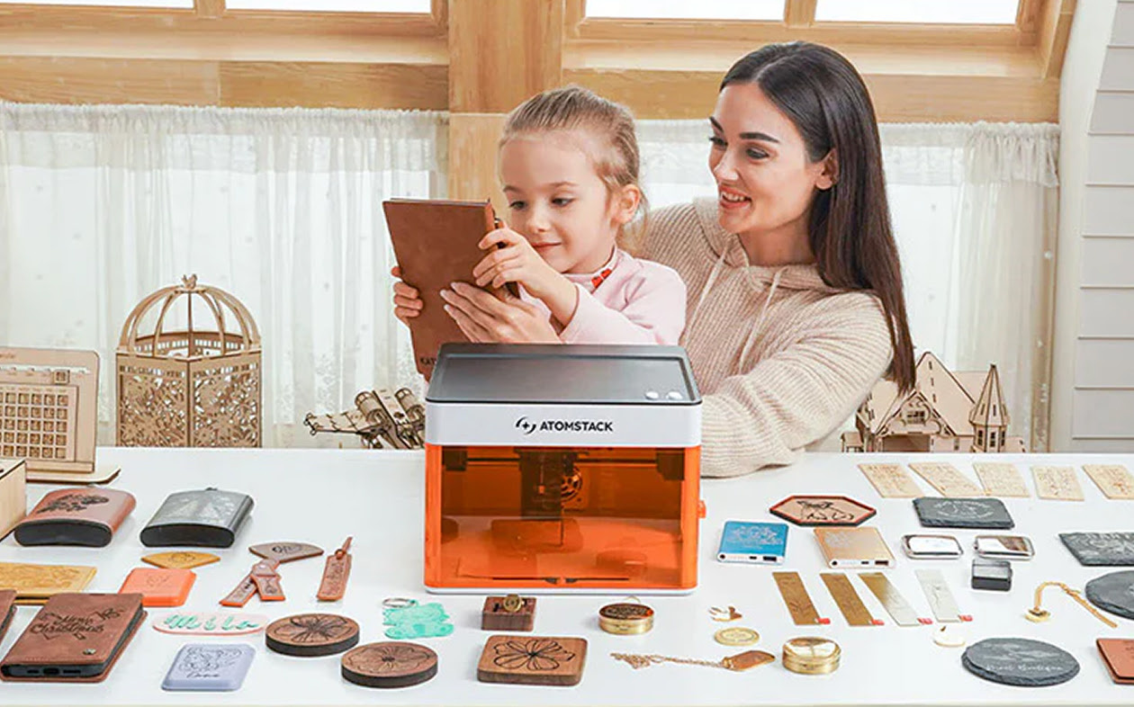 Woman sitting at a table with a child. On the table is an AtomStack P1 and a lot of laser engraved products. The child is holding a journal with leather cover.