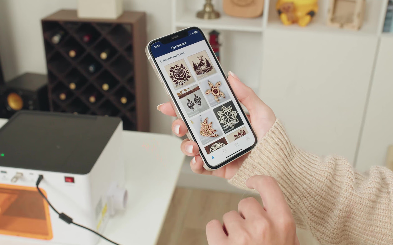 Hand holding a smartphone with AtomStack Studio opened showing the template library. In the background is an AtomStack P1 on a table.