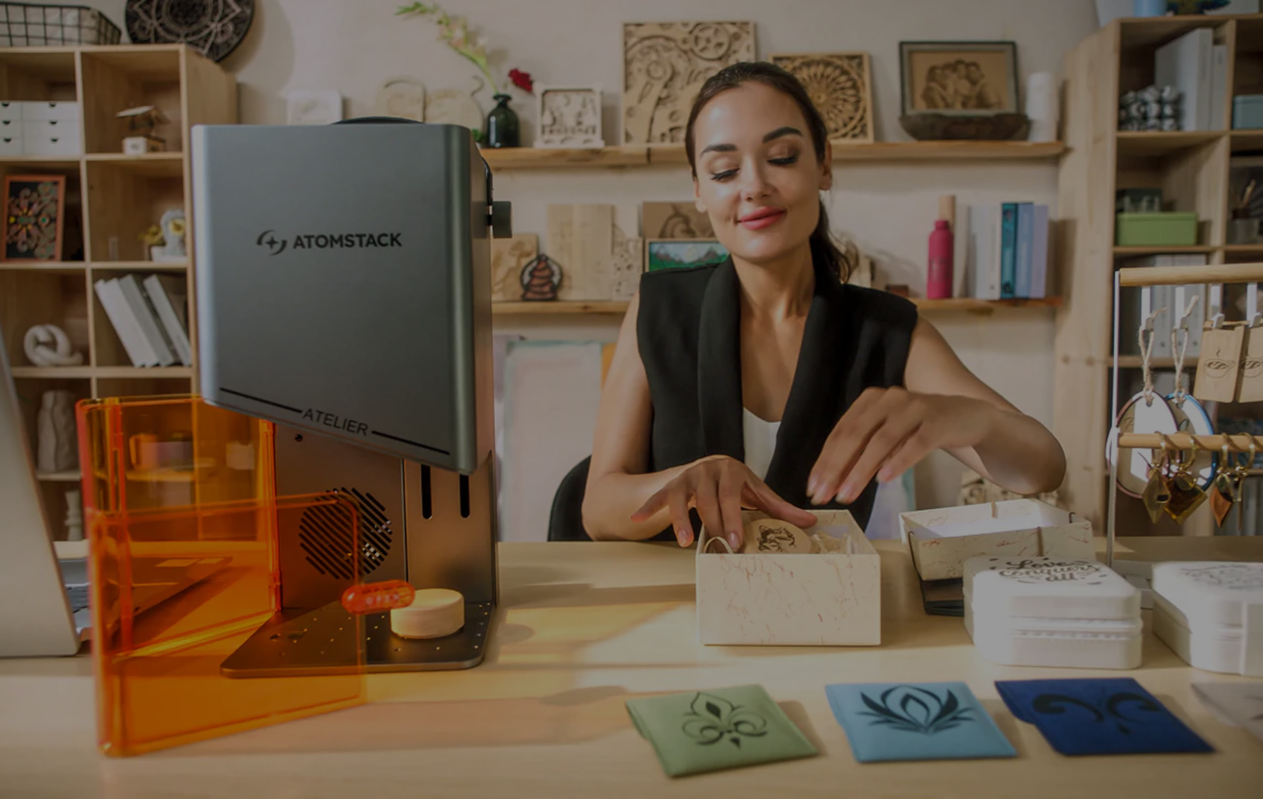 AtomStack Atelier on a desk with the door open and a wooden disk inside. A wooman is also sitting at the desk.