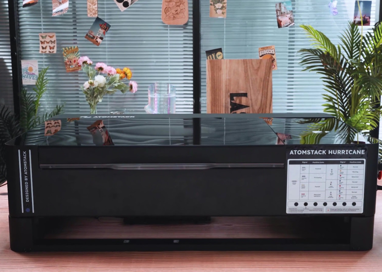 AtomStack Hurricane with the riser base installed. The laser cutter is standing on a wooden table. In the background is a window with the blinds pulled down and ferns on the left and right side. Cards mounted on strings are dangling in front of the window.