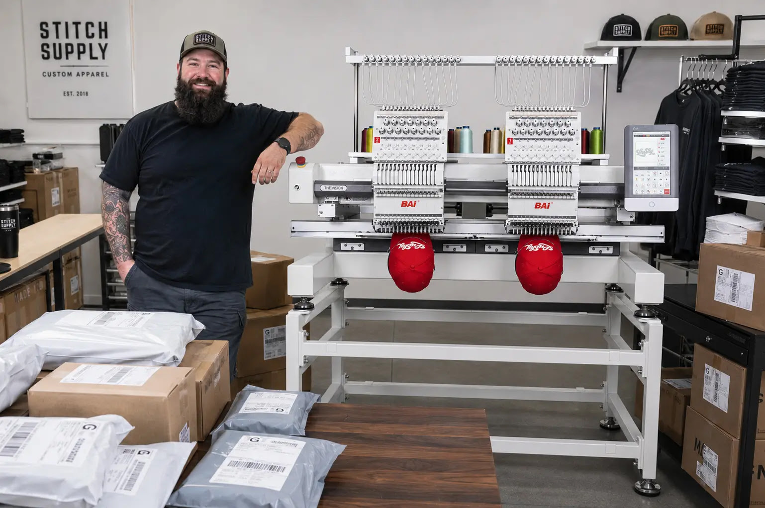Bearded operator standing by a BAi The Vision 2-Head commercial embroidery machine set up with red caps in a custom apparel studio surrounded by packaged orders
